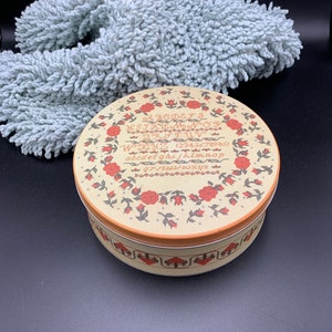May include: A round, cream-colored tin with a red and black floral design. The tin has a red and black border with a white alphabet and numbers around the edge. The tin is sitting on a black surface.