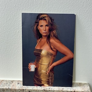 May include: A woman in a gold dress holds a glass of milk. She has a white mustache from the milk.