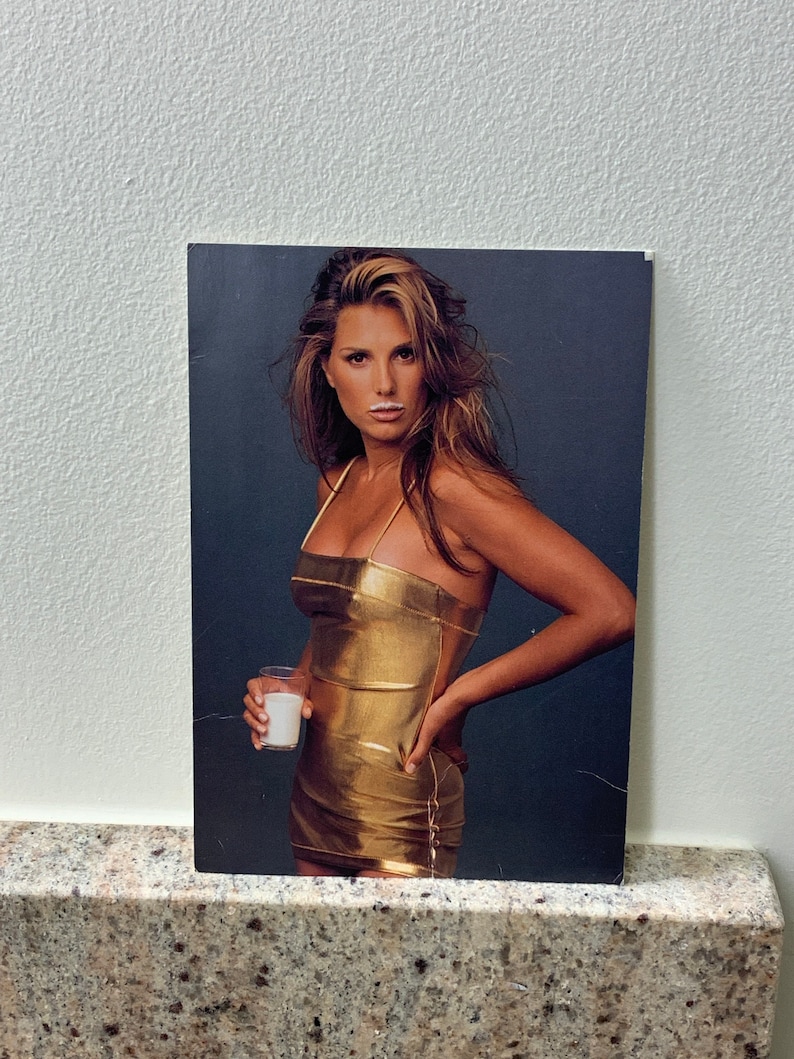 May include: A woman in a gold dress holds a glass of milk. She has a white mustache from the milk.