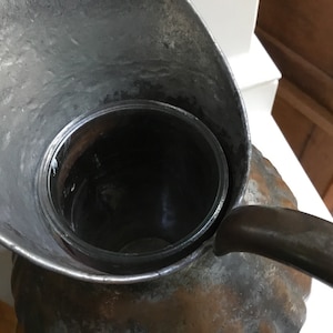 K&ouml;nnte beinhalten: Eine Vintage-Metallkanne mit Ausguss und Griff, mit einem klaren Glaseinsatz. Die Kanne hat ein verwittertes, antikes Aussehen mit sichtbaren Gebrauchsspuren. Das Design umfasst einen breiten Ausguss und einen gebogenen Griff.