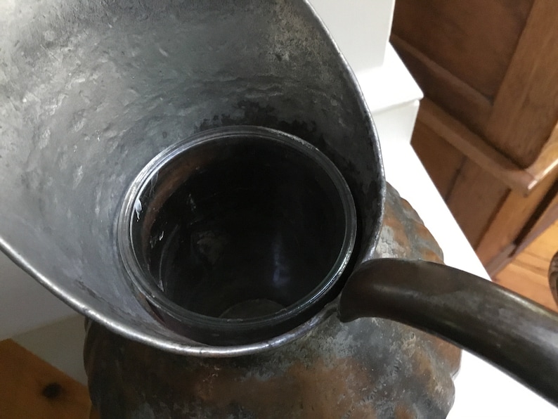 K&ouml;nnte beinhalten: Eine Vintage-Metallkanne mit Ausguss und Griff, mit einem klaren Glaseinsatz. Die Kanne hat ein verwittertes, antikes Aussehen mit sichtbaren Gebrauchsspuren. Das Design umfasst einen breiten Ausguss und einen gebogenen Griff.