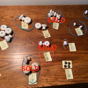 May include: Assortment of white ceramic cabinet knobs with metal accents, some are sold, others are available for purchase.  The knobs are arranged on a wooden surface with handwritten notes indicating the price and quantity.  A pile of screws is also visible in the image.