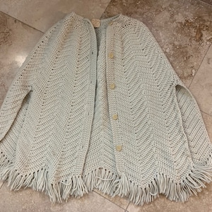 May include: A light blue crocheted poncho with a fringe border and button closures.