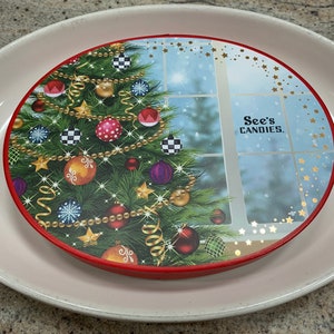 May include: A round red and gold Christmas-themed tin box with a picture of a decorated Christmas tree and a window with falling snow. The box has the text "See's CANDIES." printed on it.