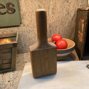 May include: A wooden potato masher with a long handle. The masher is brown and has a rectangular shape. It is sitting on a white marble cutting board.
