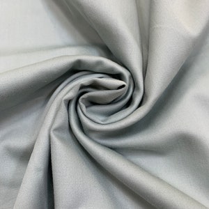 May include: A close-up of a light gray fabric with a smooth texture. The fabric is folded and draped, creating a soft and flowing look.