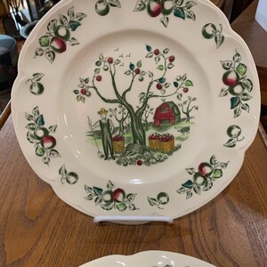 May include: Two white ceramic plates with a scalloped edge. The plates feature a green, red, and brown apple orchard scene with a man picking apples. The plates are perfect for serving a meal or displaying as decorative pieces.