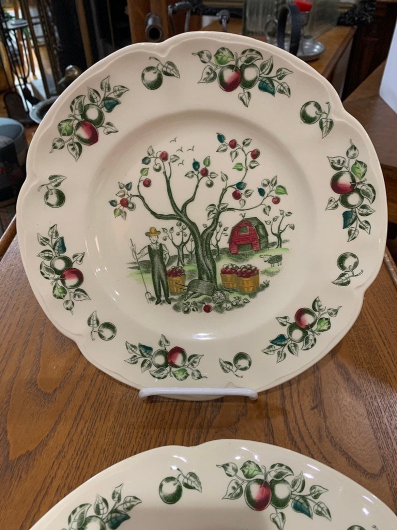 May include: Two white ceramic plates with a scalloped edge. The plates feature a green, red, and brown apple orchard scene with a man picking apples. The plates are perfect for serving a meal or displaying as decorative pieces.