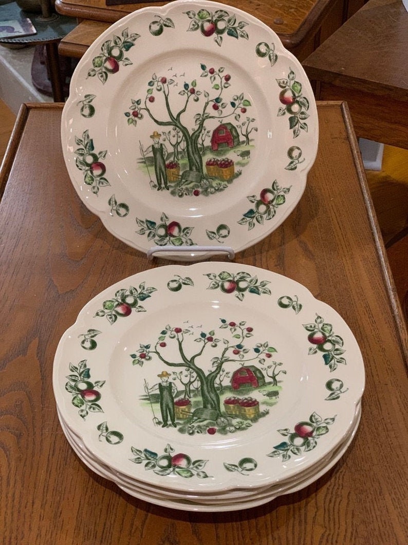 May include: A set of four white ceramic dinner plates with a scalloped edge. Each plate features a green and red apple tree with a red barn and a person picking apples. The plates are decorated with green leaves and red apples.