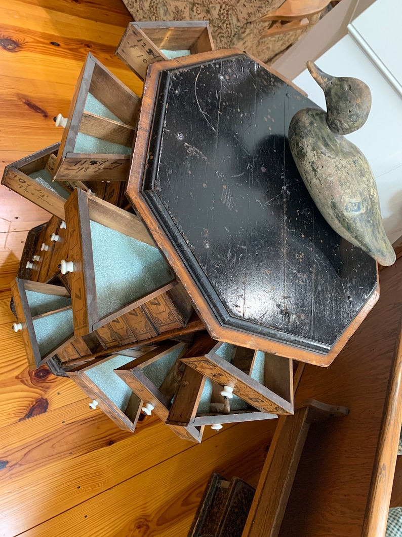 Puede incluir: Un mueble antiguo de madera con m&uacute;ltiples cajones peque&ntilde;os, cada uno con un pomo blanco. El mueble est&aacute; sujeto a un respaldo de madera negro octogonal. Un se&ntilde;uelo de pato de madera descansa sobre el respaldo.