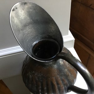 K&ouml;nnte beinhalten: Antiker Metallkrug mit breitem Ausguss und gebogenem Griff. Der Krug hat einen strukturierten, gerippten K&ouml;rper und ein dunkles, gealtertes Aussehen. Der Ausguss hat eine hellere, silberne Farbe.