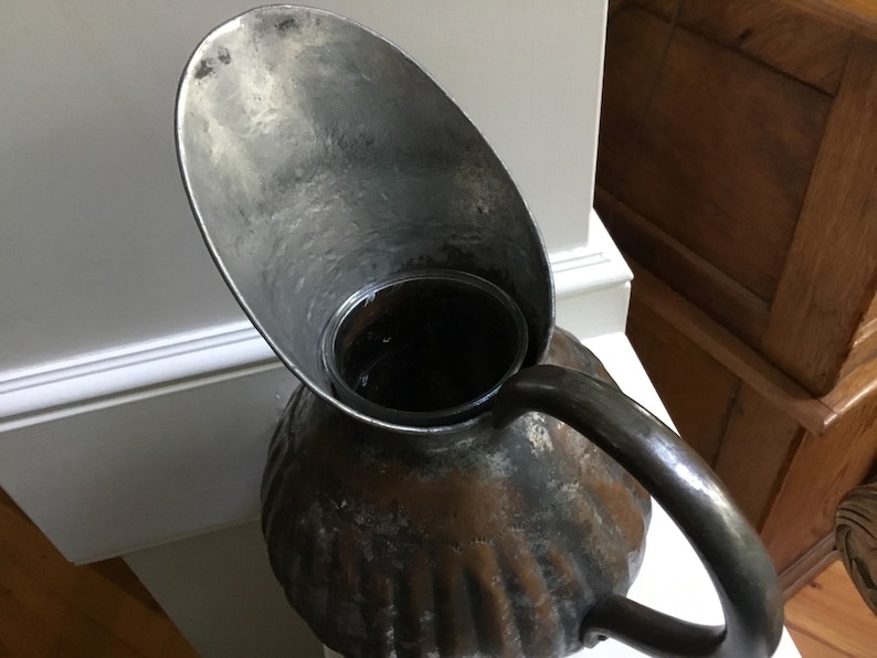 K&ouml;nnte beinhalten: Antiker Metallkrug mit breitem Ausguss und gebogenem Griff. Der Krug hat einen strukturierten, gerippten K&ouml;rper und ein dunkles, gealtertes Aussehen. Der Ausguss hat eine hellere, silberne Farbe.