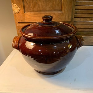 May include: A brown glazed ceramic crock with a lid and two handles. The crock is round and has a smooth, glossy finish.