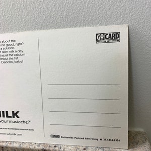 May include: A white postcard with a black and white illustration of a milk mustache. The text on the postcard reads "Milk your mustache?" and "Why Milk?" The postcard also includes the text "GoCard Nationwide Postcard Advertising 213.665.2556".