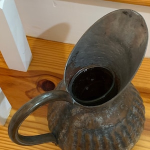K&ouml;nnte beinhalten: Ein antiker Metallkrug mit gro&szlig;em Ausguss und Griff. Der Krug hat ein strukturiertes, geripptes Design am K&ouml;rper und eine dunkle, gealterte Oberfl&auml;che. Der Griff ist gebogen und an der Seite befestigt. Der Ausguss ist breit und oval geformt.