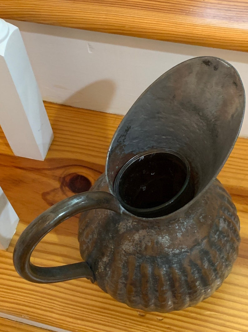 K&ouml;nnte beinhalten: Ein antiker Metallkrug mit gro&szlig;em Ausguss und Griff. Der Krug hat ein strukturiertes, geripptes Design am K&ouml;rper und eine dunkle, gealterte Oberfl&auml;che. Der Griff ist gebogen und an der Seite befestigt. Der Ausguss ist breit und oval geformt.