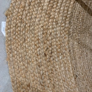 May include: A natural fiber braided rug in a light beige color. The rug is made of tightly woven strands of jute, creating a textured and durable surface.
