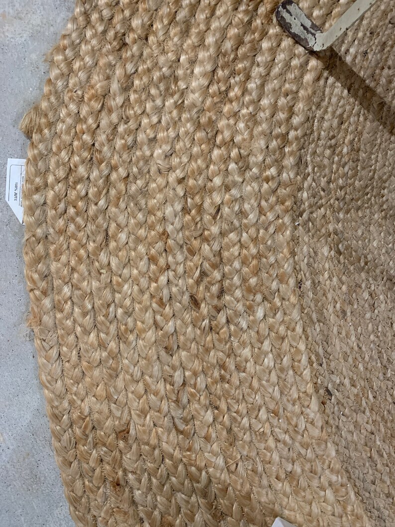 May include: A natural fiber braided rug in a light beige color. The rug is made of tightly woven strands of jute, creating a textured and durable surface.