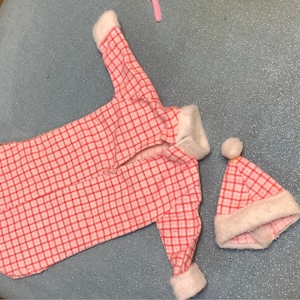 May include: A pink and white checkered doll outfit with a long-sleeved shirt and a matching hat. The outfit has white fur trim around the edges.
