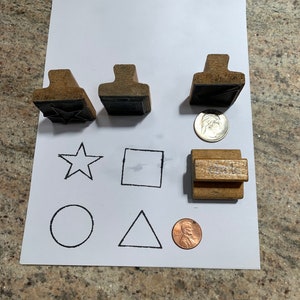 May include: Four wooden stamps with different shapes: a star, a square, a circle, and a triangle. The stamps are arranged on a white sheet of paper with the shapes drawn in black ink. There are two copper coins on the paper.