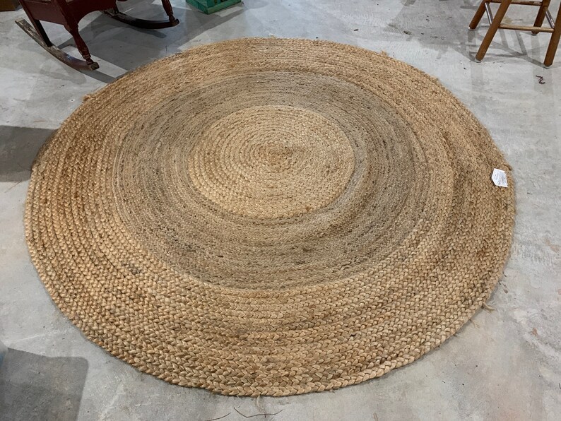 May include: A round, natural fiber rug with a woven design. The rug has a light brown color with a darker brown center.
