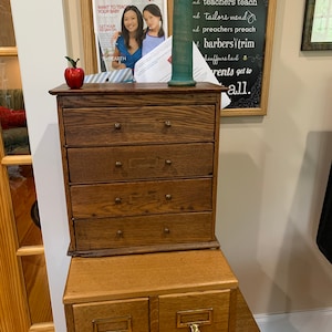 May include: A stack of two wooden drawer units. The top unit has five drawers with brass knobs. The bottom unit has two drawers with label holders. A red apple and a green vase sit on top. A magazine cover is visible.
