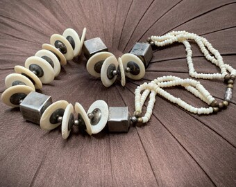 May include: A white and silver necklace with round and square beads. The necklace is made of multiple strands of small white beads and larger, round white beads with silver accents. The necklace is on a brown fabric background.