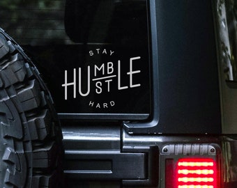 May include: A black Jeep with a white vinyl sticker on the back window that says "Stay Hustle Hard" with the word "Hustle" divided by a line with "MB" above the line and "ST" below the line. The Jeep has a red taillight.