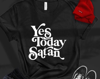 May include: Black t-shirt with white text that reads "Yes Today Satan" in a retro font style. The shirt is laid flat on a dark surface with a pair of black sneakers and blue jeans.