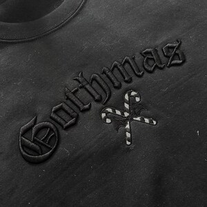 May include: Black crew neck sweatshirt with the word "Gothmaz" embroidered in gothic font. A candy cane and bat wing design is embroidered below the text. The sweatshirt is made of a soft material.