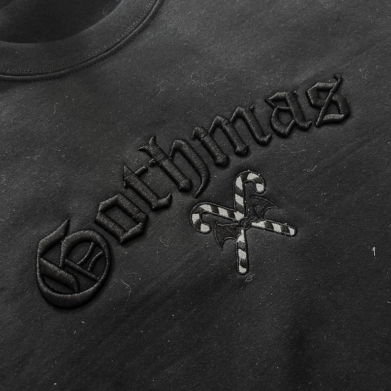 May include: Black crew neck sweatshirt with the word "Gothmaz" embroidered in gothic font. A candy cane and bat wing design is embroidered below the text. The sweatshirt is made of a soft material.