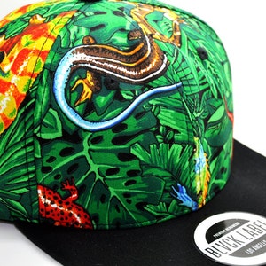 May include: A baseball cap featuring a vibrant jungle print with green leaves, a brown and blue lizard, and a red spotted frog. The cap has a solid black brim and a circular sticker on the brim that reads BLVCK LABEL PREMIUM HEADWEAR LOS ANGELES.