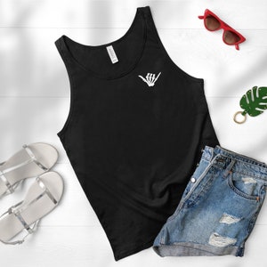 Op de afbeelding: Zwart tanktop met een witte skelethand die een shaka-teken op de voorkant maakt. Het tanktop ligt op een wit oppervlak met een paar zilveren sandalen, een rode zonnebril en een groene blaadjes sleutelhanger.