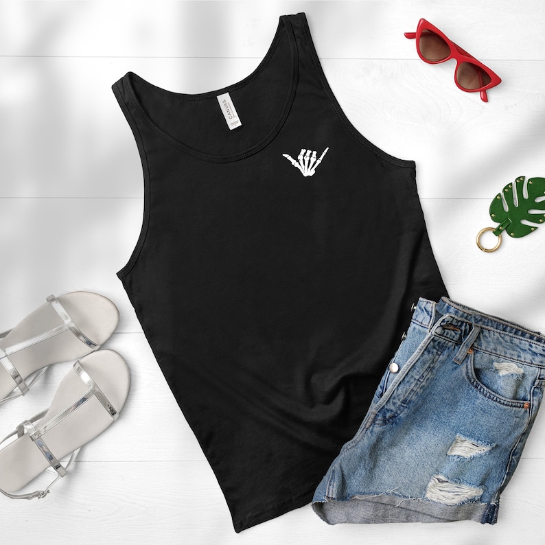 Op de afbeelding: Zwart tanktop met een witte skelethand die een shaka-teken op de voorkant maakt. Het tanktop ligt op een wit oppervlak met een paar zilveren sandalen, een rode zonnebril en een groene blaadjes sleutelhanger.