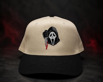 Può includere: Un cappellino da baseball beige con visiera nera, caratterizzato da un ricamo raffigurante una figura incappucciata con una maschera da fantasma bianca che impugna un coltello argentato con una goccia di sangue rosso sulla lama.
