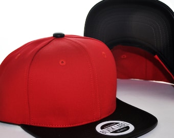May include: A red baseball cap with a black flat brim and a black button on top. The brim features a circular sticker with the text BLVCK LABEL LOS ANGELES. A second black baseball cap is positioned behind the first, showing its interior lining.