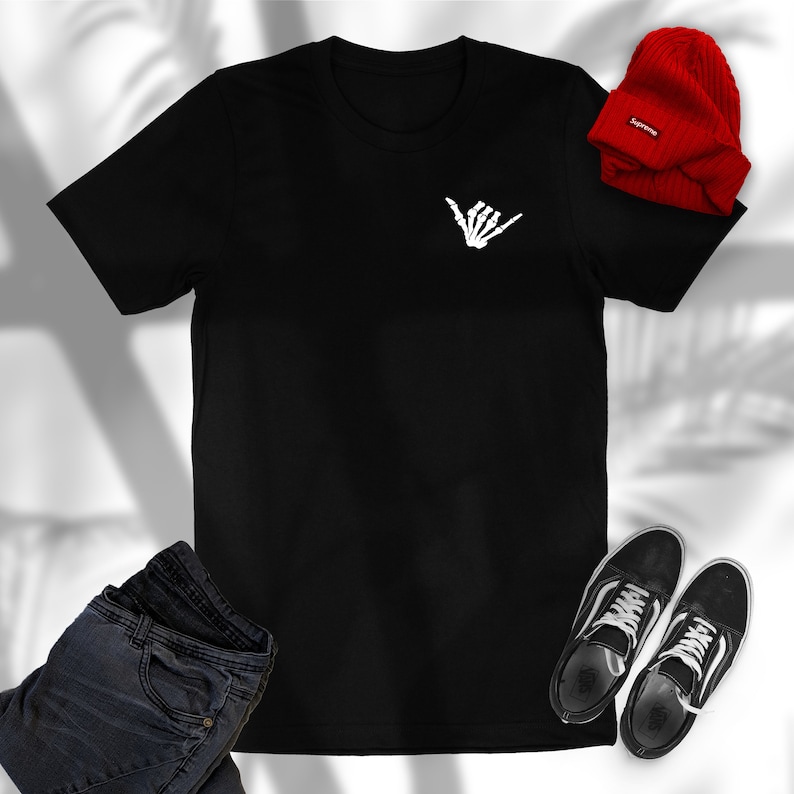 Op de afbeelding: Zwart T-shirt met een witte skelethand die een shaka-teken maakt. Het T-shirt ligt plat op een wit oppervlak met een paar zwarte sneakers en een rode beanie met het woord "Supreme" erop.