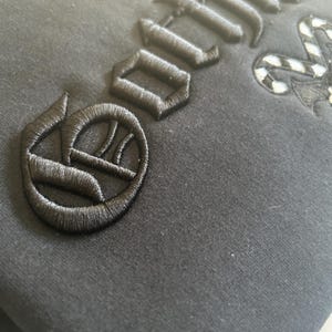 May include: Close-up of a black garment with embroidered black lettering and a decorative emblem. The stitching is detailed and raised, creating a textured effect against the fabric. The design appears to be gothic-inspired.