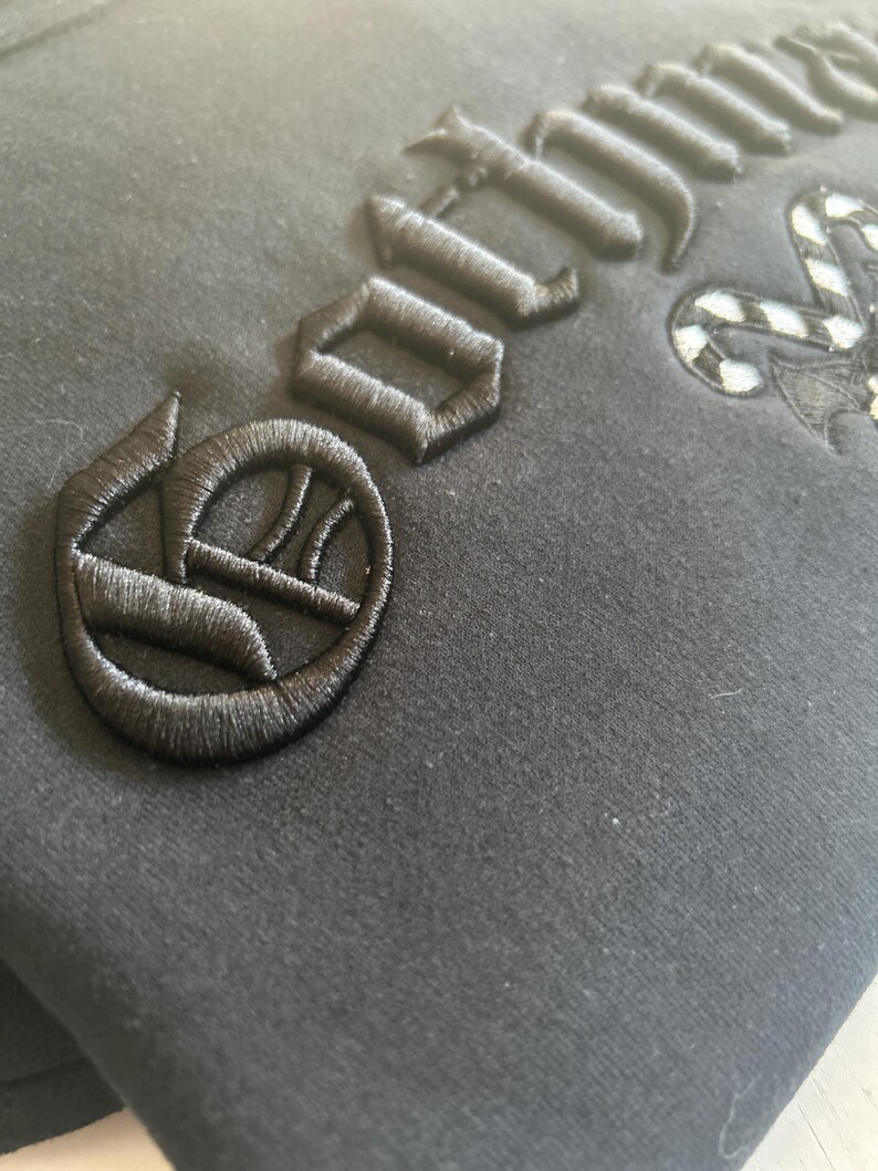 May include: Close-up of a black garment with embroidered black lettering and a decorative emblem. The stitching is detailed and raised, creating a textured effect against the fabric. The design appears to be gothic-inspired.