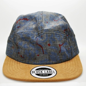 May include: A five-panel cap with a blue and gray patterned crown, accented with red paint-like splatters. The cap features a tan, curved brim and a circular label with the text "BLVCK LABEL LOS ANGELES".