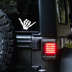 May include: A black Jeep with a white skeleton hand "hang loose" sticker on the back window. The Jeep has a red taillight with a black housing.