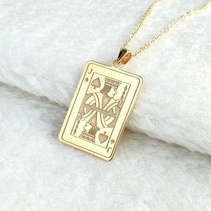 May include: Gold-coloured pendant necklace featuring a rectangular pendant with a detailed engraving of the Jack of Spades playing card. The pendant hangs from a delicate gold chain, set against a white, textured surface.