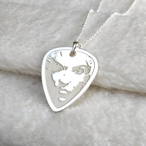 Puede incluir: Un collar con colgante plateado con un dije en forma de púa de guitarra. El dije tiene un retrato grabado detallado. La cadena del collar también es plateada. El fondo es una superficie suave, blanca y texturizada.