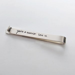 May include: Silver tie bar with the inscription "you're a warrior. love u" engraved on it.