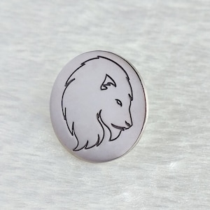 Pode incluir: Um alfinete redondo prateado com um contorno preto que representa o perfil de um urso. O urso está virado para a direita, com a cabeça e a parte superior do corpo visíveis. O alfinete está sobre um fundo branco texturizado.