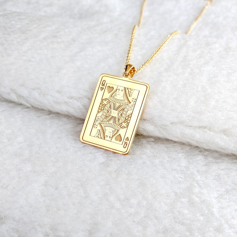May include: A gold pendant necklace with a rectangular pendant engraved with the Queen of Hearts playing card design. The pendant is suspended from a fine gold chain. The necklace is presented on a white, textured background, creating a luxurious feel.