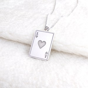 May include: A silver necklace with a pendant in the shape of a playing card. The card is the Ace of Hearts, with the heart symbol in the centre and the letter "A" at the top and bottom corners. The necklace hangs on a fine chain.