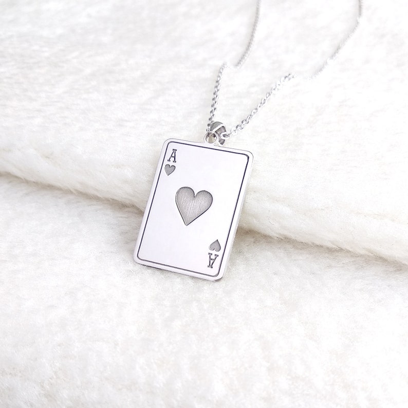 May include: A silver necklace with a pendant in the shape of a playing card. The card is the Ace of Hearts, with the heart symbol in the centre and the letter "A" at the top and bottom corners. The necklace hangs on a fine chain.