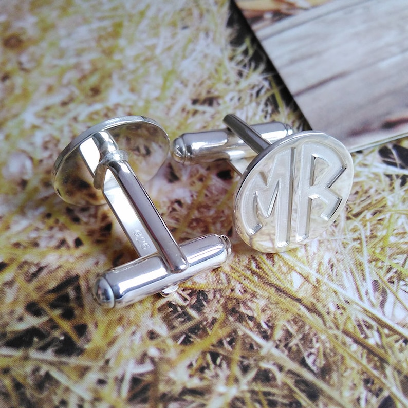 Puede incluir: Un par de gemelos de plata con un dise&ntilde;o circular. Un gemelo presenta las iniciales "MR" grabadas en la parte delantera. El otro gemelo tiene una superficie lisa.