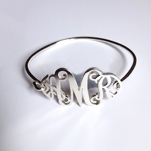 Può includere: Un braccialetto rigido in argento con un ciondolo monogramma con le lettere "MCR" in un carattere corsivo.
